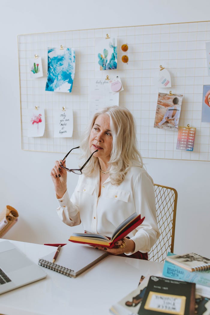 Elderly woman with glasses reading a book while seated at a desk, deep in thought.