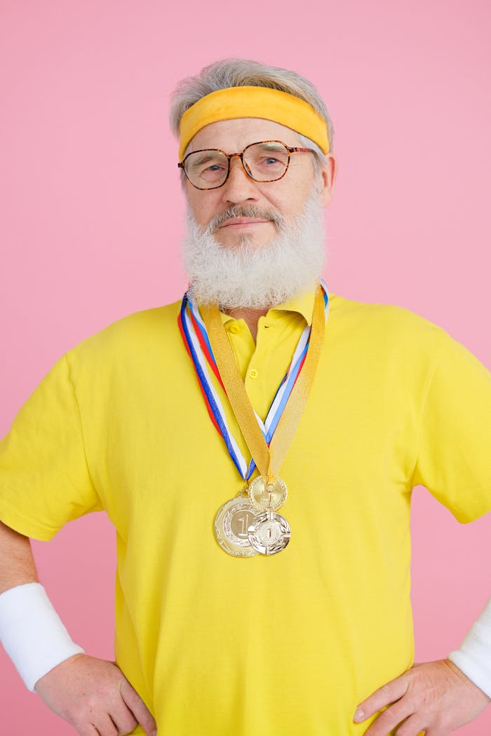 Confident senior man in yellow activewear with medals, posing on a pink background.