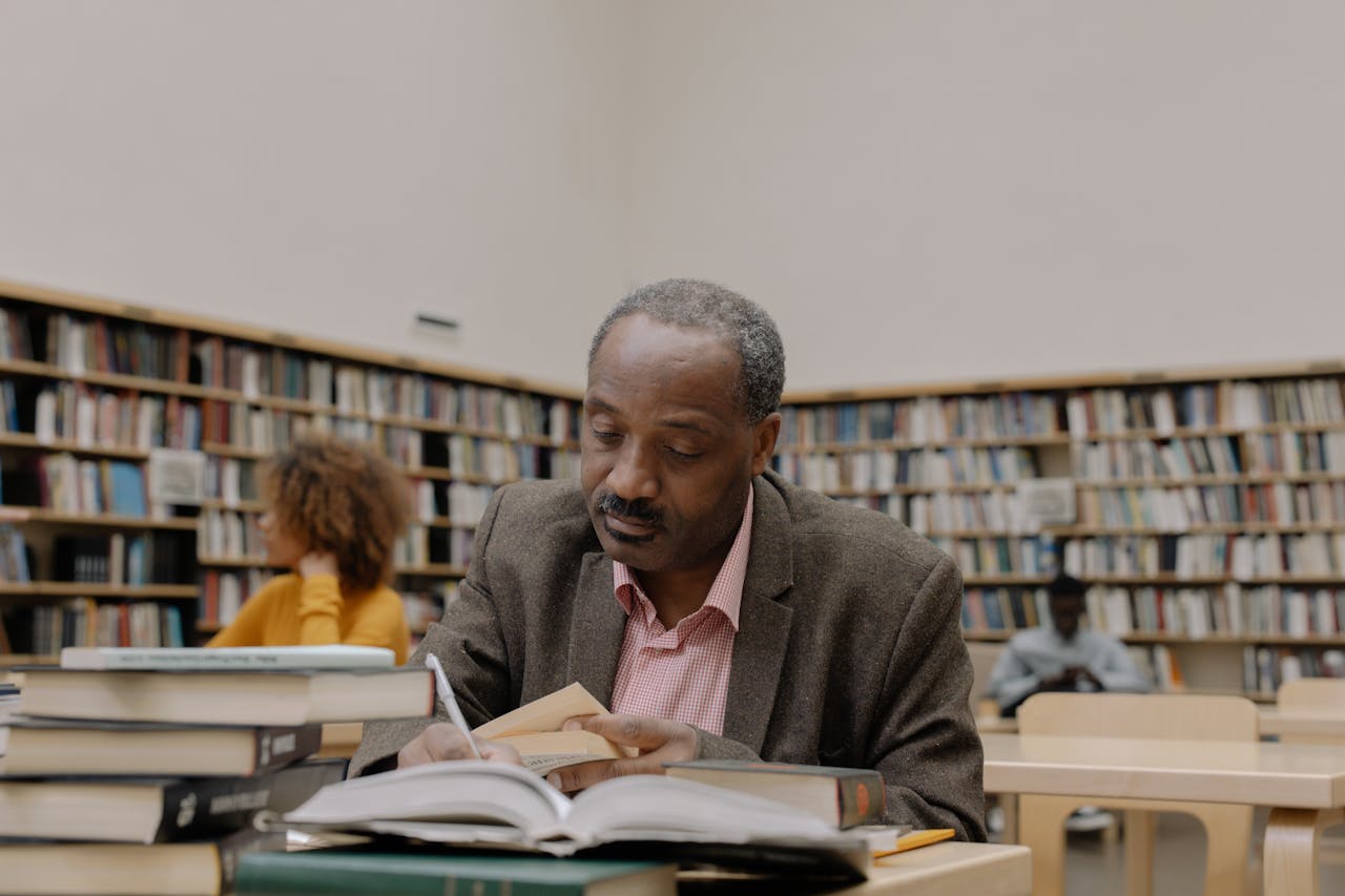 An adult man reads and studies at a desk in a public library, surrounded by bookshelves.