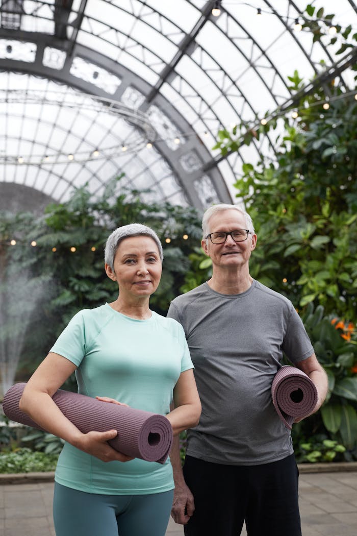 Happy senior couple holding yoga mats in a lush greenhouse setting.